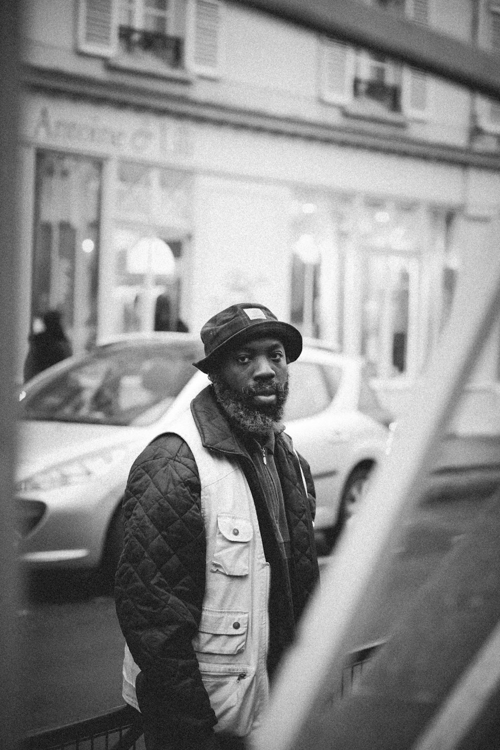 grayscale photo of man in black jacket and black hat standing near car