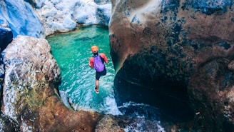 A person is preparing to jump into a clear turquoise pool of water located within a rocky gorge. Wearing a helmet and carrying gear, the individual appears ready for an adventure amidst the natural rock formations that surround the pool.