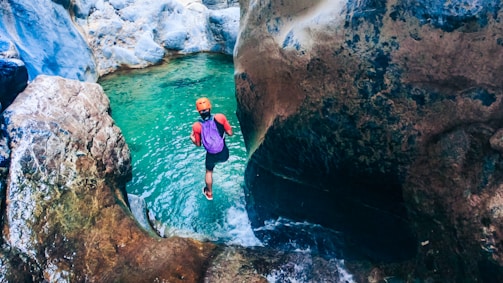 A person is preparing to jump into a clear turquoise pool of water located within a rocky gorge. Wearing a helmet and carrying gear, the individual appears ready for an adventure amidst the natural rock formations that surround the pool.