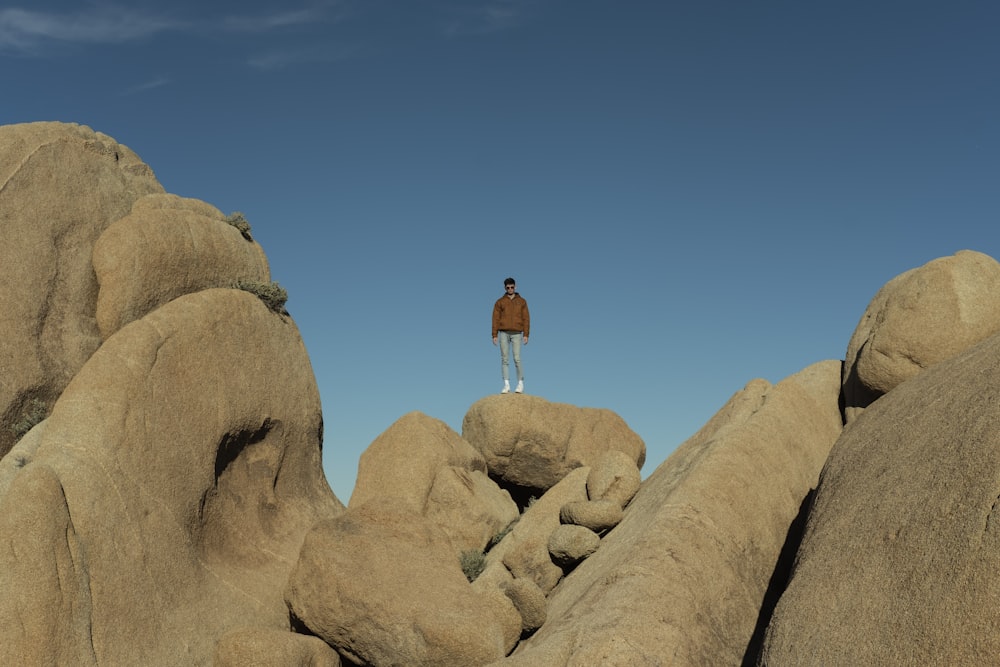 man in brown t-shirt and white shorts standing on brown rock formation under blue sky