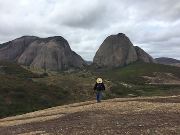 A person wearing a large hat and backpack is walking towards two large, distinctive rock formations in a rugged, green landscape under a cloudy sky.