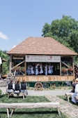 A group of people is performing a traditional dance on a wooden stage with a tiled roof, surrounded by greenery. They wear traditional costumes, including white dresses and hats. Onlookers sit on wooden benches, watching the performance in an outdoor setting. A banner hangs from the stage.