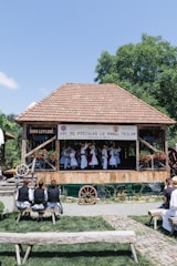 A group of people is performing a traditional dance on a wooden stage with a tiled roof, surrounded by greenery. They wear traditional costumes, including white dresses and hats. Onlookers sit on wooden benches, watching the performance in an outdoor setting. A banner hangs from the stage.