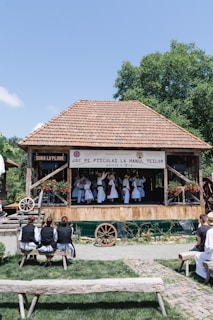 A group of people is performing a traditional dance on a wooden stage with a tiled roof, surrounded by greenery. They wear traditional costumes, including white dresses and hats. Onlookers sit on wooden benches, watching the performance in an outdoor setting. A banner hangs from the stage.