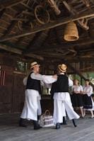 A group of people dressed in traditional folk costumes are in a wooden structure. Two men wearing hats are dancing, while two women in skirts watch. Above them, wooden beams and wheels are part of the rustic ceiling design. A woven basket hangs from the ceiling, and a bag with traditional patterns lies on the floor.