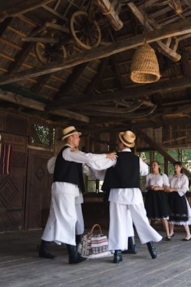 A group of people dressed in traditional folk costumes are in a wooden structure. Two men wearing hats are dancing, while two women in skirts watch. Above them, wooden beams and wheels are part of the rustic ceiling design. A woven basket hangs from the ceiling, and a bag with traditional patterns lies on the floor.