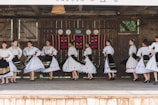 A group of people dressed in traditional folk costumes, performing a dance on a wooden stage. The costumes are predominantly white with ornate embroidered patterns, featuring floral and geometric designs. Black skirts with colorful accents and lace detailing are part of the outfits. The stage background includes rustic wooden walls with decorative plates and a colorful woven tapestry.