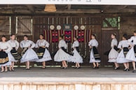 A group of people dressed in traditional folk costumes, performing a dance on a wooden stage. The costumes are predominantly white with ornate embroidered patterns, featuring floral and geometric designs. Black skirts with colorful accents and lace detailing are part of the outfits. The stage background includes rustic wooden walls with decorative plates and a colorful woven tapestry.