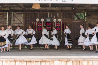 A group of people dressed in traditional folk costumes, performing a dance on a wooden stage. The costumes are predominantly white with ornate embroidered patterns, featuring floral and geometric designs. Black skirts with colorful accents and lace detailing are part of the outfits. The stage background includes rustic wooden walls with decorative plates and a colorful woven tapestry.
