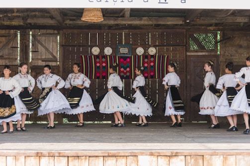 A group of people dressed in traditional folk costumes, performing a dance on a wooden stage. The costumes are predominantly white with ornate embroidered patterns, featuring floral and geometric designs. Black skirts with colorful accents and lace detailing are part of the outfits. The stage background includes rustic wooden walls with decorative plates and a colorful woven tapestry.