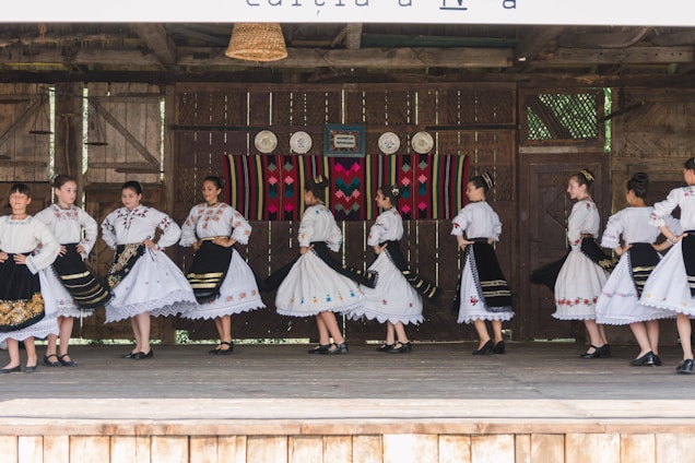 A group of people dressed in traditional folk costumes, performing a dance on a wooden stage. The costumes are predominantly white with ornate embroidered patterns, featuring floral and geometric designs. Black skirts with colorful accents and lace detailing are part of the outfits. The stage background includes rustic wooden walls with decorative plates and a colorful woven tapestry.