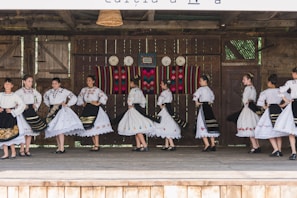 A group of people dressed in traditional folk costumes, performing a dance on a wooden stage. The costumes are predominantly white with ornate embroidered patterns, featuring floral and geometric designs. Black skirts with colorful accents and lace detailing are part of the outfits. The stage background includes rustic wooden walls with decorative plates and a colorful woven tapestry.