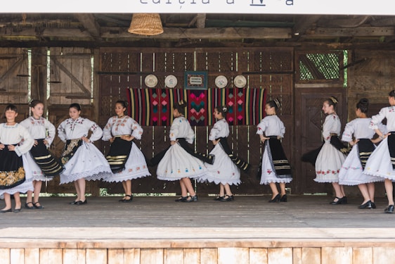 A group of people dressed in traditional folk costumes, performing a dance on a wooden stage. The costumes are predominantly white with ornate embroidered patterns, featuring floral and geometric designs. Black skirts with colorful accents and lace detailing are part of the outfits. The stage background includes rustic wooden walls with decorative plates and a colorful woven tapestry.