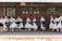 A group of people dressed in traditional folk costumes, performing a dance on a wooden stage. The costumes are predominantly white with ornate embroidered patterns, featuring floral and geometric designs. Black skirts with colorful accents and lace detailing are part of the outfits. The stage background includes rustic wooden walls with decorative plates and a colorful woven tapestry.