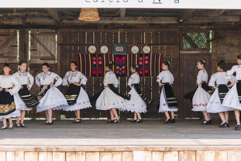 A group of people dressed in traditional folk costumes, performing a dance on a wooden stage. The costumes are predominantly white with ornate embroidered patterns, featuring floral and geometric designs. Black skirts with colorful accents and lace detailing are part of the outfits. The stage background includes rustic wooden walls with decorative plates and a colorful woven tapestry.