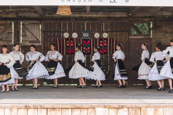 A group of people dressed in traditional folk costumes, performing a dance on a wooden stage. The costumes are predominantly white with ornate embroidered patterns, featuring floral and geometric designs. Black skirts with colorful accents and lace detailing are part of the outfits. The stage background includes rustic wooden walls with decorative plates and a colorful woven tapestry.