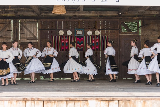 A group of people dressed in traditional folk costumes, performing a dance on a wooden stage. The costumes are predominantly white with ornate embroidered patterns, featuring floral and geometric designs. Black skirts with colorful accents and lace detailing are part of the outfits. The stage background includes rustic wooden walls with decorative plates and a colorful woven tapestry.