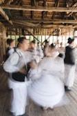 A group of people are engaged in traditional dance, wearing traditional or folkloric clothing. The setting is a rustic wooden building with large windows, and the movement of the dancers creates a dynamic and blurred effect.