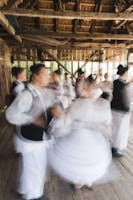 A group of people are engaged in traditional dance, wearing traditional or folkloric clothing. The setting is a rustic wooden building with large windows, and the movement of the dancers creates a dynamic and blurred effect.