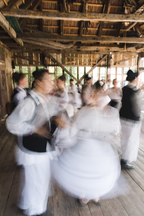 A group of people are engaged in traditional dance, wearing traditional or folkloric clothing. The setting is a rustic wooden building with large windows, and the movement of the dancers creates a dynamic and blurred effect.