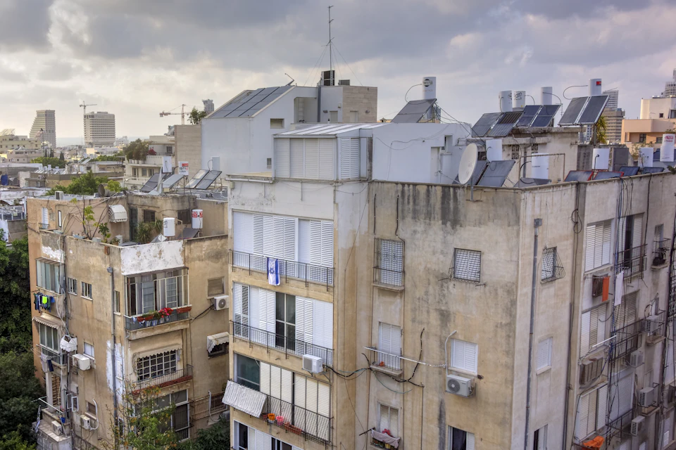 The image presents an aerial view of a cityscape, dominated by tall buildings with white and beige facades. The buildings are densely packed together, creating a sense of urban density. Some of these structures have solar panels installed on their roofs, indicating the city's commitment to sustainable energy practices. The sky above is filled with clouds, casting a soft light over the scene below.