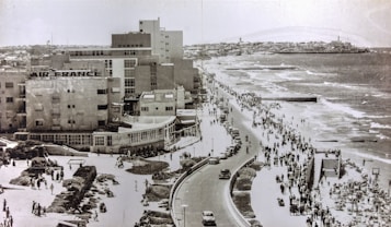 A vintage photograph capturing a bustling coastal city scene with a large beachfront promenade. The shore is lined with people walking and engaging in various activities, while cars are traveling along the road nearby. Several large buildings are situated to the left, one prominently marked with 'Air France'. The ocean waves gently lap against the beach, creating a lively and inviting atmosphere.