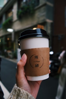 A close-up of a hand holding a travel coffee cup with a secure lid, against a city street background