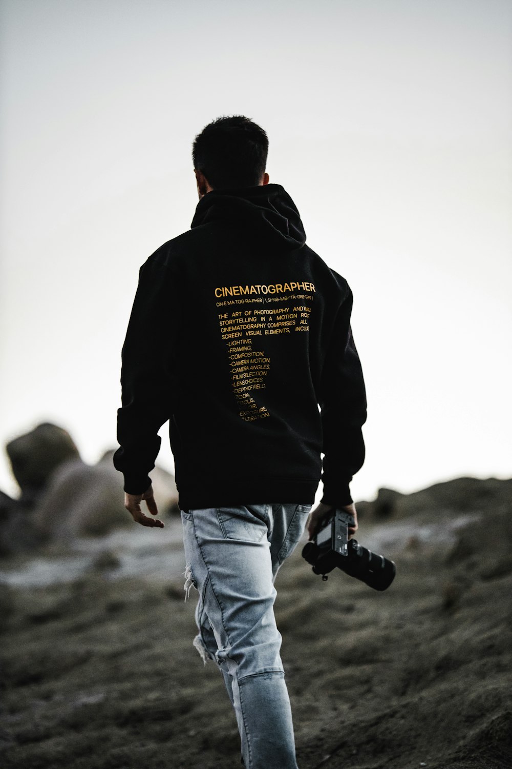 Hombre con sudadera con capucha negra y jeans de mezclilla blancos sosteniendo una cámara DSLR negra