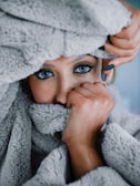 A person with striking blue eyes is partially covered by a soft, gray, fuzzy blanket. The individual is holding the blanket close to their face, creating a sense of coziness and warmth. Their hand, with neatly manicured nails, is visible gently grasping the blanket.