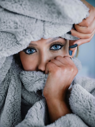 A person with striking blue eyes is partially covered by a soft, gray, fuzzy blanket. The individual is holding the blanket close to their face, creating a sense of coziness and warmth. Their hand, with neatly manicured nails, is visible gently grasping the blanket.