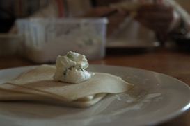 A close-up view of a plate with thin pastry sheets topped with a dollop of soft cheese. The background shows a blurred container, possibly containing more cheese, and an out-of-focus person handling food.