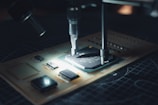 A close-up view of a microchip on a circuit board being worked on with precision instruments. The scene is illuminated, highlighting the intricate electronic components and the fine details of the circuit.