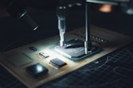 A close-up view of a microchip on a circuit board being worked on with precision instruments. The scene is illuminated, highlighting the intricate electronic components and the fine details of the circuit.