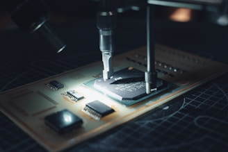A close-up view of a microchip on a circuit board being worked on with precision instruments. The scene is illuminated, highlighting the intricate electronic components and the fine details of the circuit.