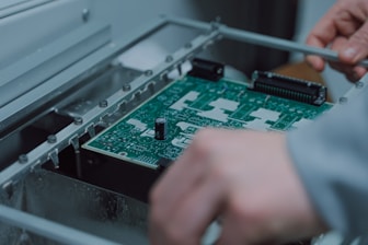 A person is assembling or inspecting a green printed circuit board (PCB) with various components mounted on it. The setting appears to be an industrial or technical environment.