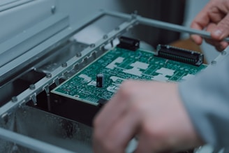 A person is assembling or inspecting a green printed circuit board (PCB) with various components mounted on it. The setting appears to be an industrial or technical environment.