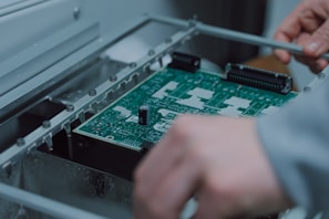 A person is assembling or inspecting a green printed circuit board (PCB) with various components mounted on it. The setting appears to be an industrial or technical environment.