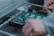 A person is assembling or inspecting a green printed circuit board (PCB) with various components mounted on it. The setting appears to be an industrial or technical environment.