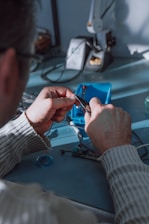 A person uses pliers to work on a small electronic component on a table. There is a soldering station and other tools in the background. The environment appears to be a workshop or electronics lab.