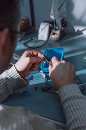 A person uses pliers to work on a small electronic component on a table. There is a soldering station and other tools in the background. The environment appears to be a workshop or electronics lab.