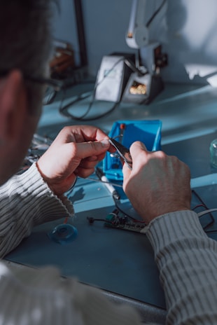 A person uses pliers to work on a small electronic component on a table. There is a soldering station and other tools in the background. The environment appears to be a workshop or electronics lab.