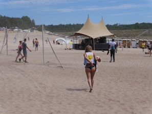 A sandy beach with several people walking and enjoying the sun. In the foreground, a person wearing a swimsuit and carrying a bag walks towards a large tented structure. The background features additional tents, people, and a forested area on the horizon.