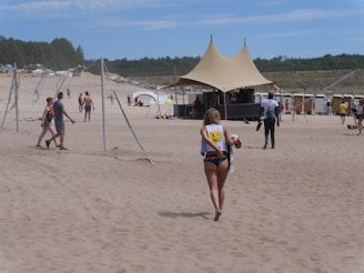 A sandy beach with several people walking and enjoying the sun. In the foreground, a person wearing a swimsuit and carrying a bag walks towards a large tented structure. The background features additional tents, people, and a forested area on the horizon.
