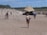 A sandy beach with several people walking and enjoying the sun. In the foreground, a person wearing a swimsuit and carrying a bag walks towards a large tented structure. The background features additional tents, people, and a forested area on the horizon.
