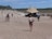 A sandy beach with several people walking and enjoying the sun. In the foreground, a person wearing a swimsuit and carrying a bag walks towards a large tented structure. The background features additional tents, people, and a forested area on the horizon.