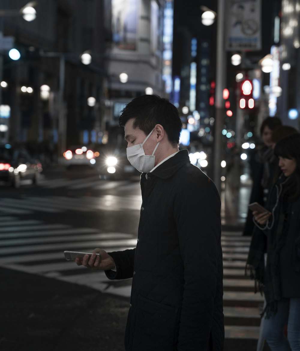 Mann in schwarzer Anzugjacke mit weißer Gesichtsmaske, der nachts auf der Fußgängerzone steht