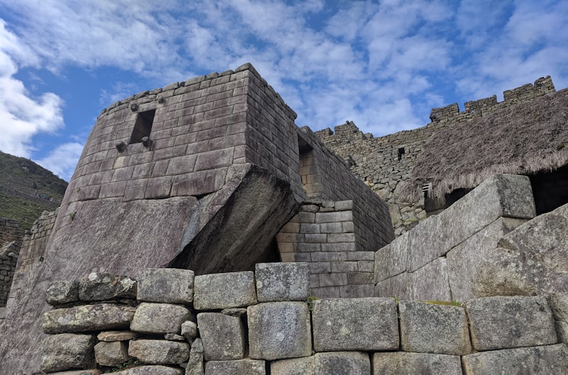 An ancient stone structure consisting of rectangular blocks meticulously arranged to form walls and a trapezoidal window, set against a partly cloudy sky. The construction features a thatched roof section and an inclined stone slab connecting to a larger wall. The architecture is characterized by its precise, interlocking design suggestive of historical significance.