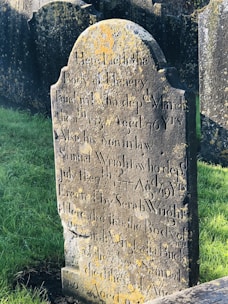 An old, weathered gravestone is covered with moss and lichen, displaying engraved text detailing the names and dates of those buried there. The stone is situated among other similar gravestones in a grassy cemetery. The lighting accentuates the texture of the stone and the growths on its surface.