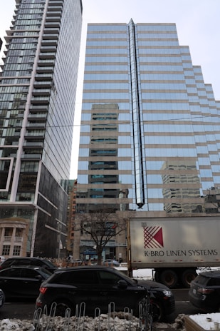 Two tall modern glass skyscrapers with reflective surfaces stand side by side, capturing the cityscape around them. A large truck with the text K-BRO Linen Systems is parked in the foreground, and several cars are lined along the street. There is snow on the ground and a row of locked bicycles, adding an urban winter setting.