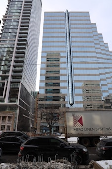 Two tall modern glass skyscrapers with reflective surfaces stand side by side, capturing the cityscape around them. A large truck with the text K-BRO Linen Systems is parked in the foreground, and several cars are lined along the street. There is snow on the ground and a row of locked bicycles, adding an urban winter setting.